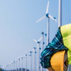 Woman engineer in uniform and holding yellow safety helmet with standing and checking wind turbine power