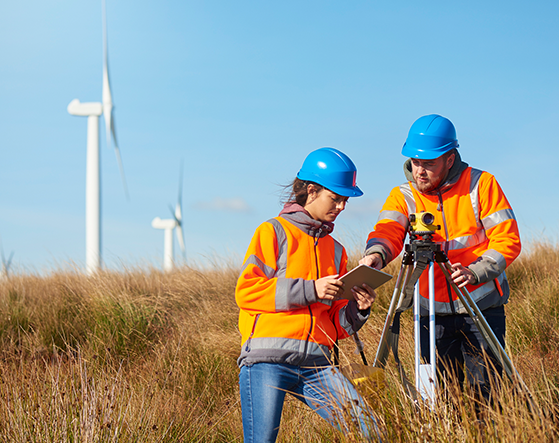 Two people in a field with hard hats and a windfarm behind them.