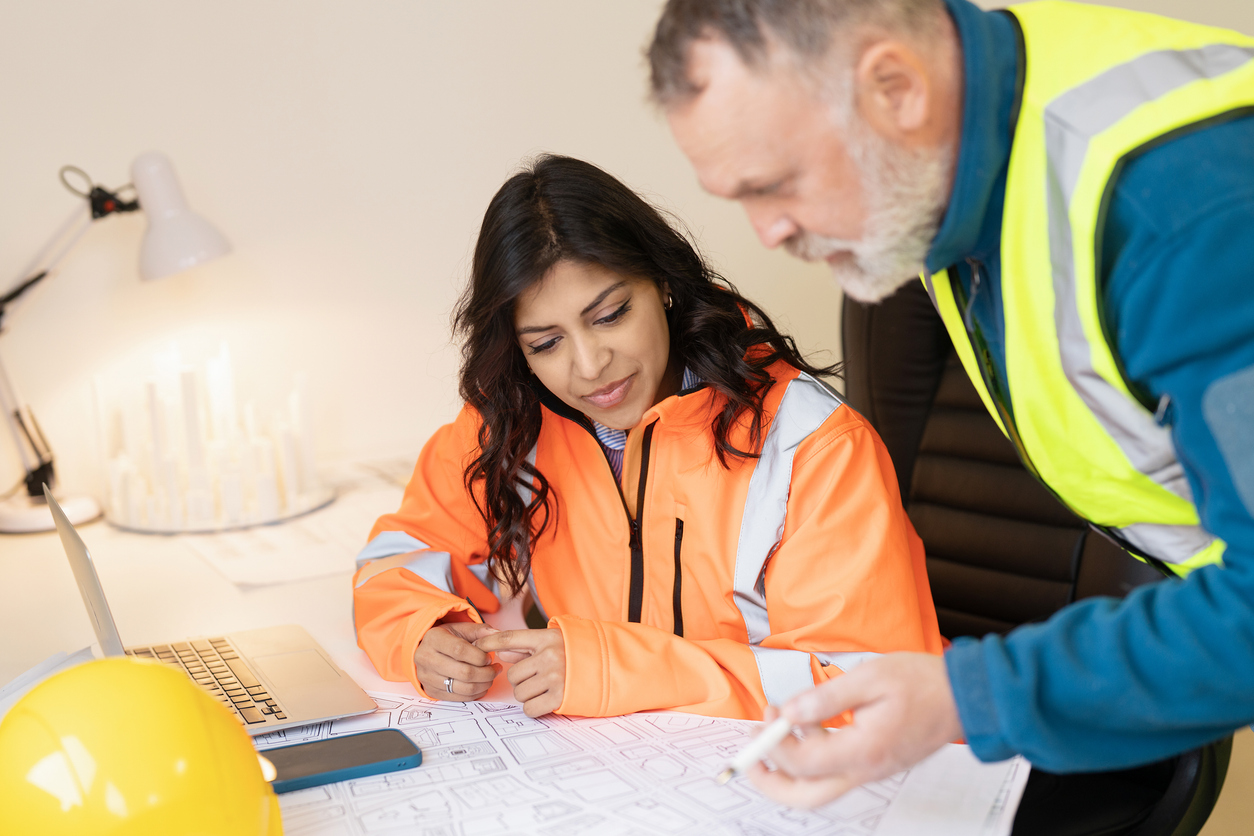 Two engineers focus on detailed construction plans within a well-lit office. One wears a safety vest and both examine designs on a table alongside a laptop and yellow helmet.