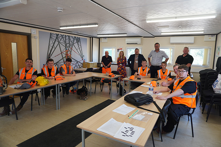 Apprentices wearing high-visibility vests attend a classroom-based training session, seated around tables with instructors and staff standing at the back of the room