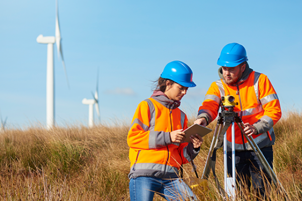 windfarm-trainee-559×443 Two people in a field with hard hats and a windfarm behind them.
